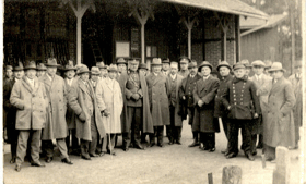 Skupina mužů před skladem, někteří v uniformě (pohled)